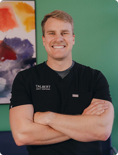 Talbert Family Dentistry | Dentist in black scrubs with 'Talbert Family Dentistry' logo, smiling with arms crossed in front of colorful abstract painting on green wall.