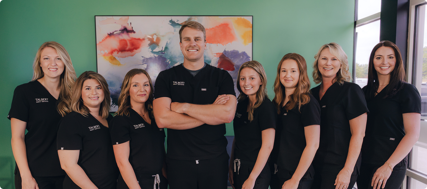 Talbert Family Dentistry | Group of dental team members in black scrubs standing together in front of green wall with colorful abstract artwork, smiling at camera.